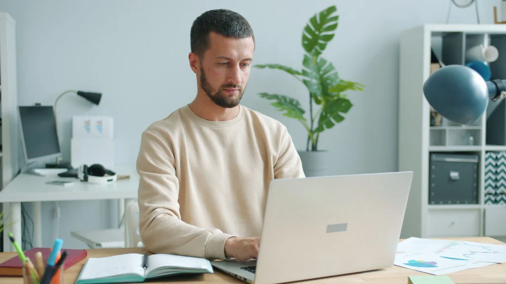Homem usa notebook em mesa de madeira cercado por materiais e equipamentos para home office modernos.