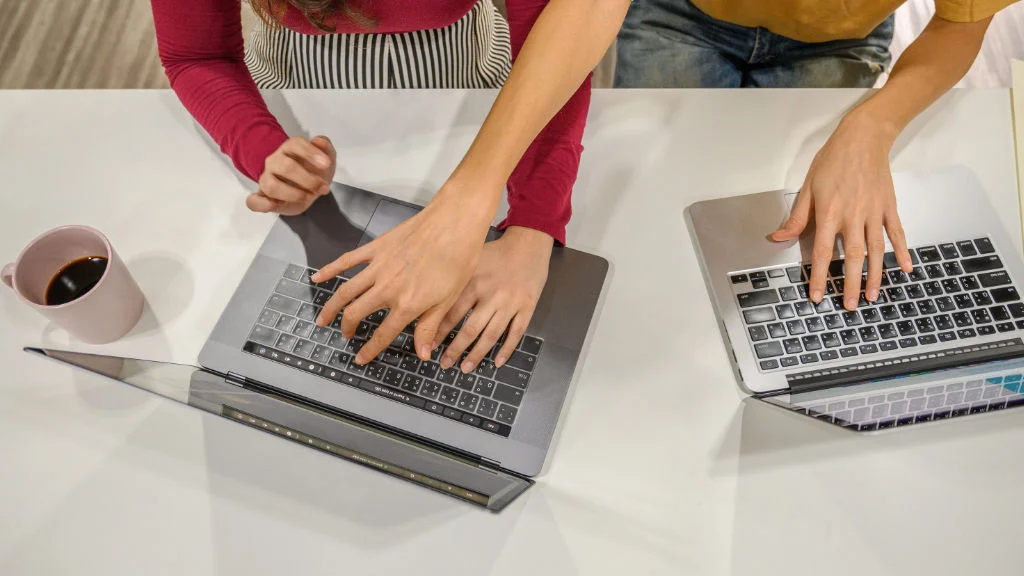 Mão de colega apontando para o teclado de outro durante sessão de trabalho colaborativo.