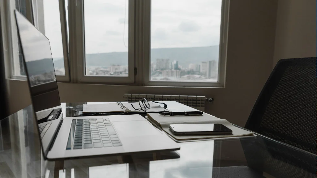 Notebook, celular, óculos e caderno organizados em mesa de vidro para um home office organizado.