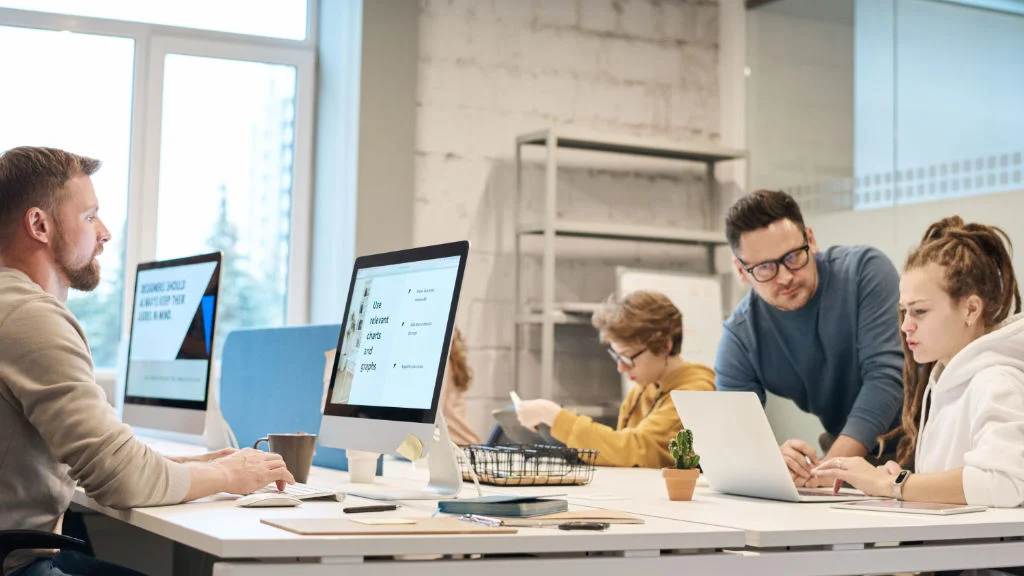 Pessoas colaborando em mesa com computadores e notebooks.