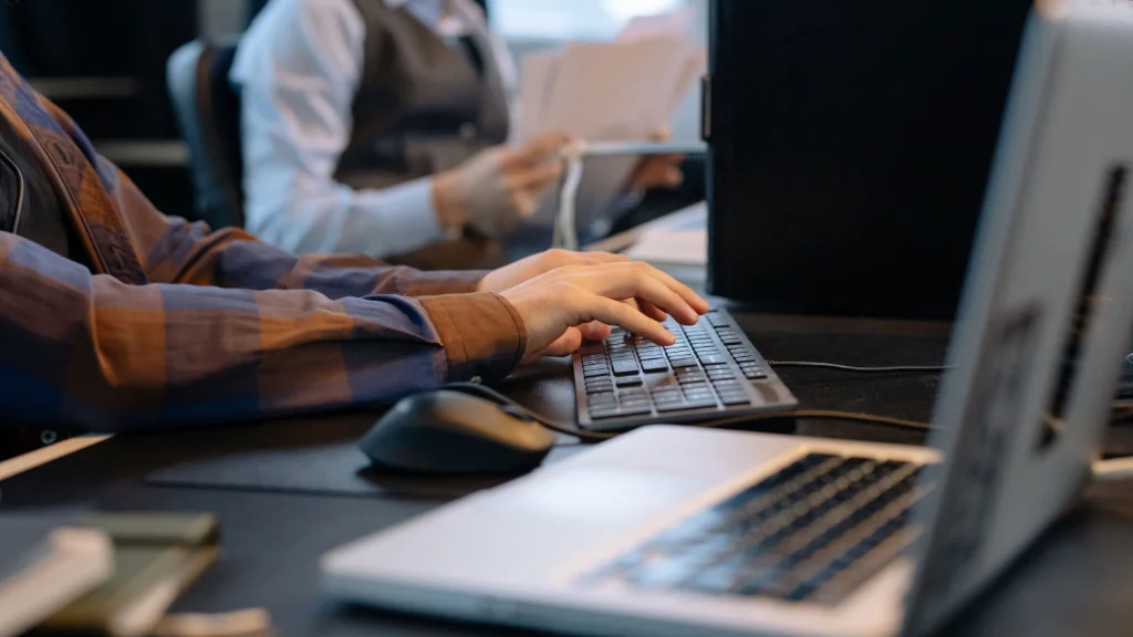 Pessoa digitando em um teclado preto, ao lado de notebook e computador, sobre a mesa em um escritório.