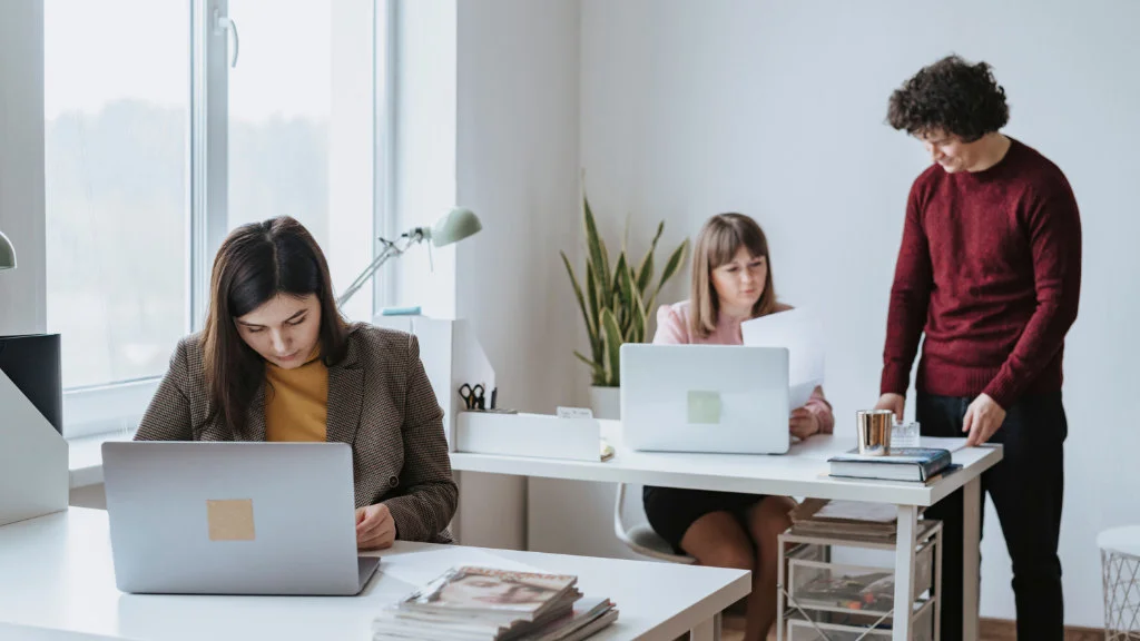 Três profissionais trabalhando em um ambiente de trabalho moderno e bem iluminado com notebooks e plantas.