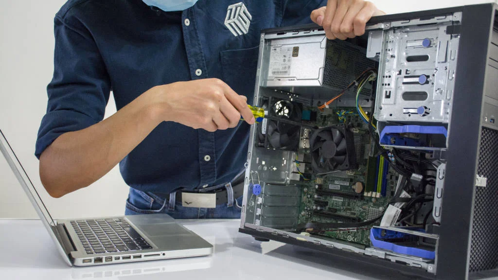 Profissional ajusta componentes de um computador aberto ao lado de notebook durante processo de gestão de equipamentos de TI.