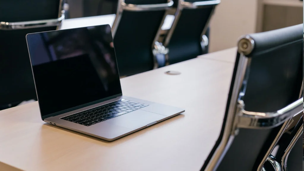 Notebook aberto sobre mesa de madeira clara em uma sala de reunião corporativa com cadeiras pretas.