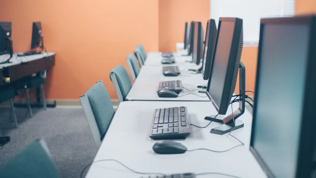 Estação de trabalho em laboratório de informática equipado com computadores alinhados em bancadas.