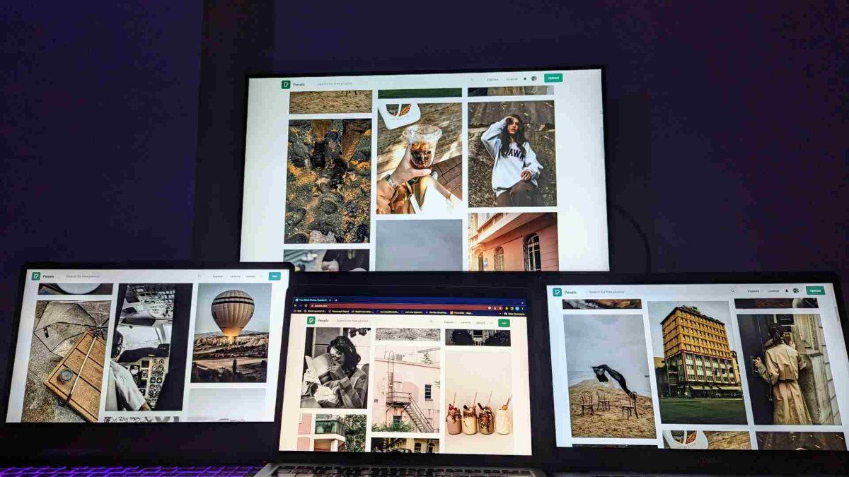 Quatro telas, incluindo notebook e PC para trabalhar, exibindo um banco de imagens em um ambiente com iluminação escura.